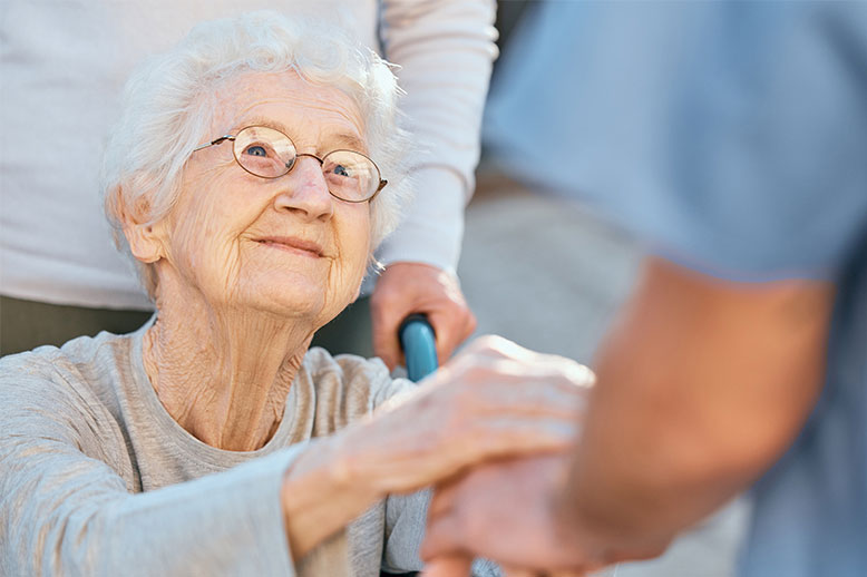 Eine ältere Frau mit Brille, die sich an der Hand einer anderen Person festhält und Hilfe oder Unterstützung erhält.